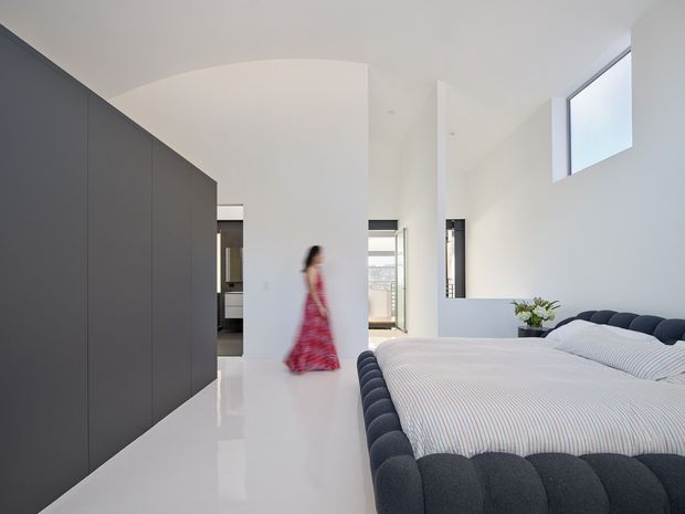 Bright contemporary bedroom with a large upholstered bed featuring a dark scalloped frame and headboard, crisp striped bedding, extensive dark built-in cabinetry along the left wall, and glossy white flooring and walls. A high horizontal window and narrow glazed openings bring in daylight, while a blurred figure adds scale.