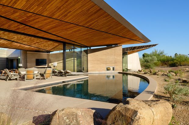 Exterior view of a contemporary single-storey residence with broad cantilevered timber soffits, extensive floor-to-ceiling glazing, and warm horizontal timber-clad walls. A curved swimming pool borders a paved outdoor living area with seating and an outdoor television, while desert-style landscaping and large natural boulders surround the property.