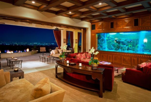 A luxurious living room opening onto a nighttime terrace, with extensive timber built-in cabinetry, a large illuminated aquarium, exposed dark timber ceiling beams, rich red upholstered seating, and a substantial dark wood coffee table. Floor-to-ceiling openings with curtains provide views of city lights, while warm layered lighting creates an opulent, dramatic atmosphere.