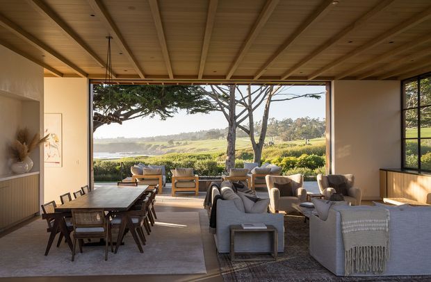 Warm, open-plan living and dining area with extensive floor-to-ceiling glazing opening toward a landscaped rural or vineyard-like setting. The room features a timber-lined exposed-beam ceiling, light neutral walls, a long dining table with timber chairs, and a relaxed arrangement of upholstered sofas and armchairs. Large openings create strong indoor–outdoor connection and expansive views.