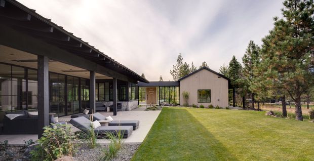 A single-storey contemporary residence in a wooded setting, viewed from a landscaped lawn. The home combines a long, low pavilion with extensive floor-to-ceiling glazing, dark exposed roof eaves, slender posts, and an adjoining simple gabled-volume wing with pale vertical cladding. A covered outdoor lounge with sofas and armchairs opens onto a paved terrace, with planting beds and a broad lawn in the foreground.