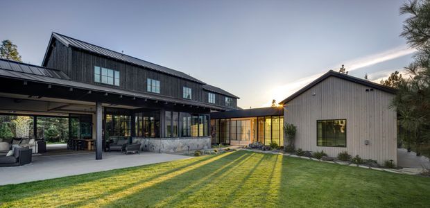 Exterior view of a contemporary multi-building residence arranged around a landscaped lawn. The main structure has dark vertical cladding, a steep metal gable roof, extensive black-framed glazing, and a covered outdoor living area. A lighter gabled wing is connected by a glazed link, creating a courtyard-like composition. The image is photographed at golden hour with long shadows across the lawn.