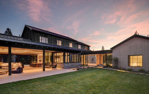 A dusk exterior view of a contemporary multi-wing residence arranged around a landscaped courtyard. The home features dark vertical timber cladding, grey metal roofing, extensive black-framed glazing, a covered outdoor living terrace, and a lighter metal-clad secondary wing. Warm interior and exterior lighting creates an inviting atmosphere.