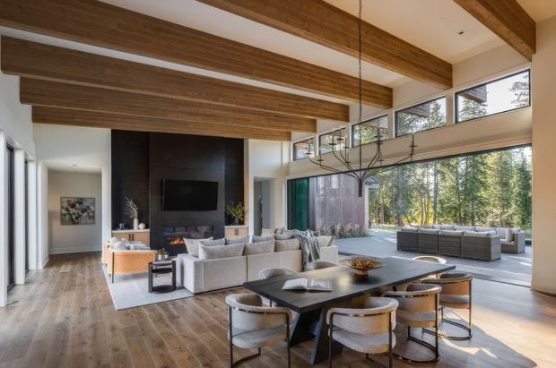 A spacious open-plan living and dining room with exposed timber ceiling beams, warm timber flooring, a large dark feature wall with fireplace and television, and expansive floor-to-ceiling glazing opening toward a furnished outdoor terrace and wooded setting. A large sectional sofa and dining table define the contemporary interior, while a sculptural linear pendant hangs above the dining area.