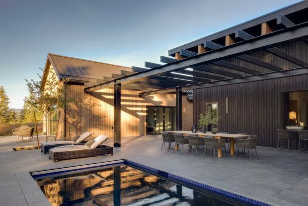 A contemporary residential outdoor entertaining area with a covered black pergola and exposed roof framing, positioned beside a reflective swimming pool. The space includes an outdoor dining table with chairs, a lounge seating area, dark vertical wall cladding, timber-lined soffits, large openings, and warm low-angle sunlight. The image presents a refined modern exterior with strong indoor-outdoor connection and expansive paved terraces.