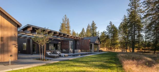 Exterior view of a contemporary, low-profile residential house set within a wooded landscape. The design features dark vertical cladding, warm timber wall panels, broad glazing, deep roof overhangs, and a paved terrace with outdoor seating and a long narrow water feature or pool. The house appears largely single-storey with layered, shallow-pitched roof forms and strong indoor-outdoor connections.