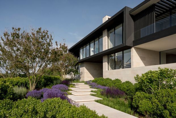 Exterior view of a contemporary multi-level residence with strong horizontal lines, expansive floor-to-ceiling glazing, dark projecting upper volumes, pale masonry walls, and a screened balcony or terrace. A broad concrete-paved path and steps lead through a formal landscaped garden with clipped shrubs, ornamental grasses, and purple flowering planting.