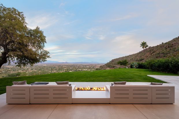 A landscaped outdoor terrace overlooking a broad valley, with a contemporary built-in outdoor lounge arranged around a long linear fire feature. The scene includes pale tiled paving, low-profile sectional seating, manicured lawn, hedging, hillside terrain, and distant mountain views under a soft evening sky.