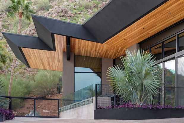 Contemporary angular residential exterior with dramatic folded black metal roof forms, warm timber soffits and wall cladding, dark framed glazing, and a raised entry terrace with glass and metal balustrades. The house is set against a rocky, planted hillside and features integrated landscaping along the facade.