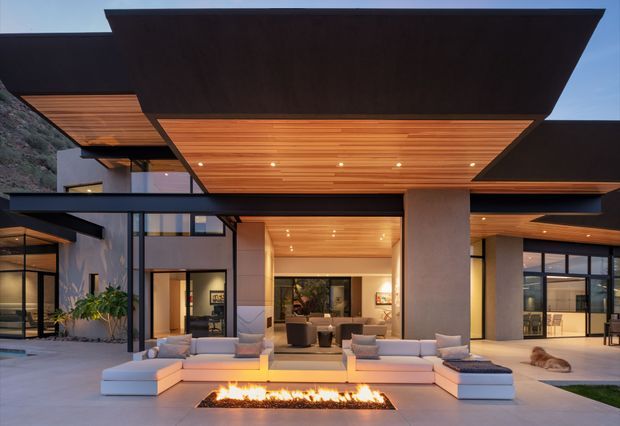 Exterior architectural photograph of a large contemporary residence at dusk, viewed from a landscaped terrace. The home features broad flat projecting roof planes, extensive dark-framed glazing, warm timber soffits with recessed lighting, pale rendered walls, and an outdoor lounge arranged around a long linear fire feature. Interior living spaces are visible through the open central facade, with a rocky hillside at the side.