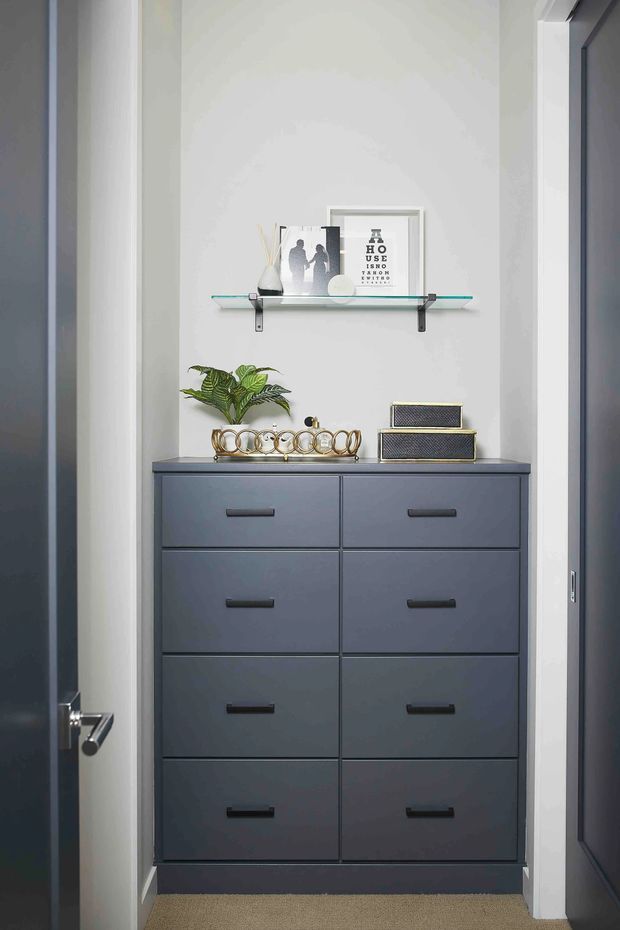 A narrow, tall built-in storage alcove or entryway is shown between dark doors. It contains a dark charcoal-grey chest of drawers with multiple flat-front drawers and black horizontal pulls, topped with decorative objects, a potted plant, and stacked boxes. A slim glass display shelf with metal brackets is mounted above against a white wall. The space has neutral finishes, carpet flooring, white trim, and a clean, orderly appearance.