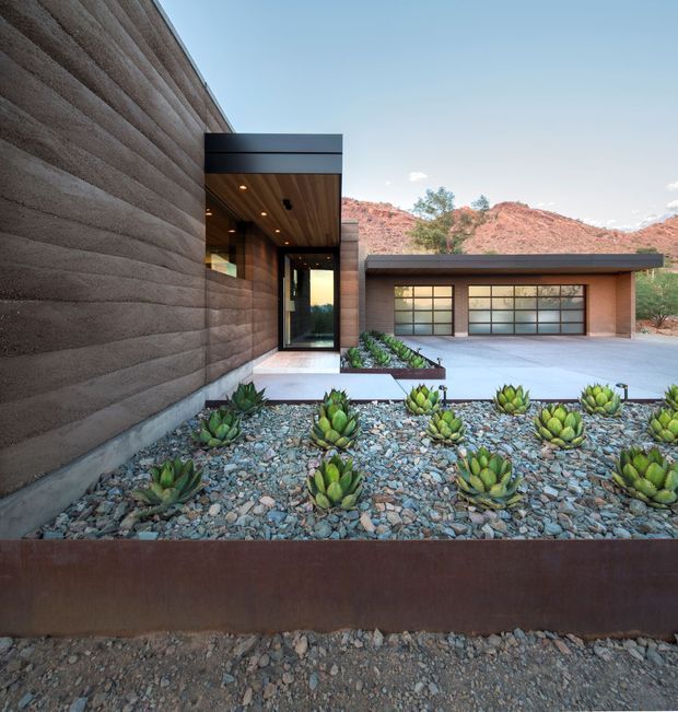 Modern single-storey desert residence with heavily textured earth-toned exterior walls, dark metal roof and trim, and expansive gridded garage-style glazing. A recessed entry is sheltered beneath a timber-lined canopy, while landscaped gravel beds with sculptural succulents lead toward the building. Red rocky desert hills and sparse vegetation form the background.