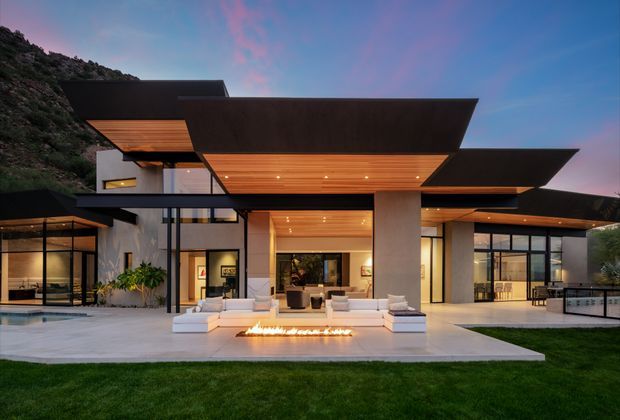 Twilight exterior view of a large contemporary hillside residence with dramatic projecting flat roof planes, extensive floor-to-ceiling glazing, exposed timber soffits, pale rendered walls, and a broad paved terrace. A linear outdoor fire feature and built-in seating occupy the foreground, with landscaped lawn and a steep vegetated hillside behind.