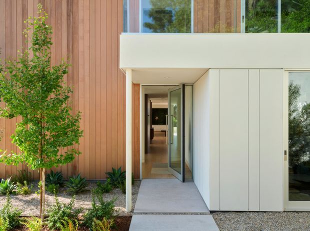A modern residential entrance with a projecting flat-roofed upper volume, pale smooth wall panels, full-height glazing, and a recessed glazed front door. Warm vertical timber cladding forms the adjoining facade, contrasting with the light facade and concrete entry path. Low-maintenance landscaped planting and gravel border the entry.