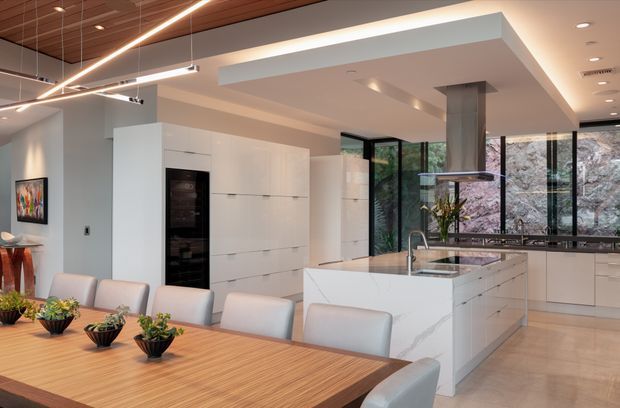 A spacious open-plan kitchen and dining area with a long light-wood dining table and upholstered chairs in the foreground. The contemporary white kitchen features extensive handleless cabinetry, a large marble-look island with integrated sink, a dark built-in appliance column, and a sleek suspended rangehood. Full-height black-framed glazing overlooks trees, while a timber slatted ceiling and linear pendant lighting add warmth and definition.