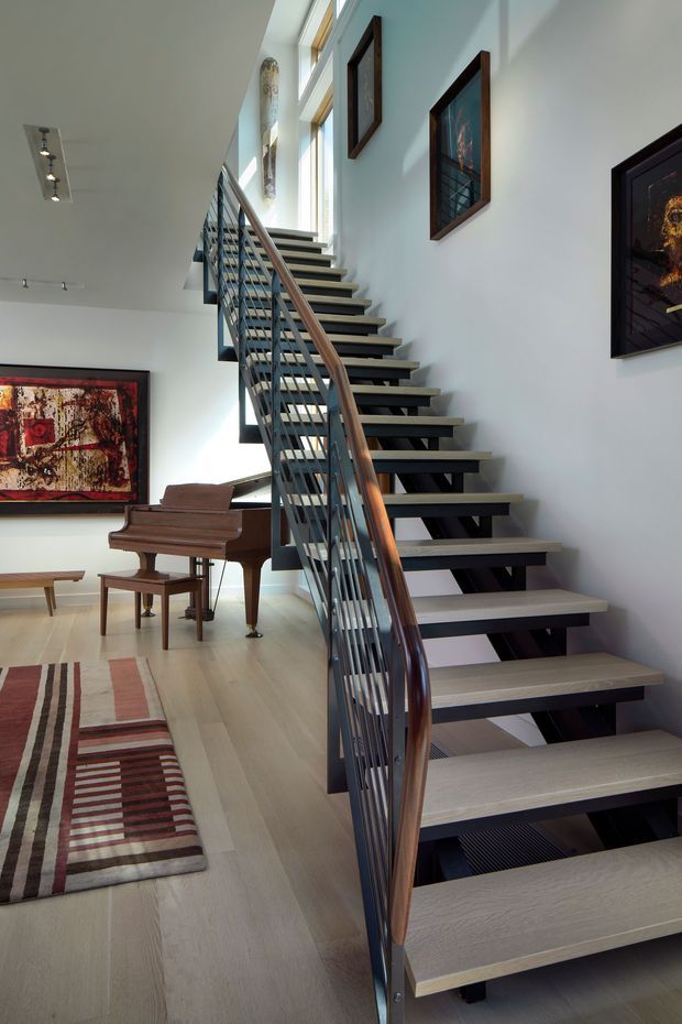 A bright, double-height interior stairway with an open-riser staircase, pale timber treads, dark metal stringers and slender metal balusters, and a curved timber handrail. The stair rises alongside tall vertical glazing and white walls displaying framed artwork. A polished grand piano, bench, patterned rug, and low timber table occupy the adjoining living area.
