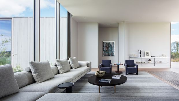 A bright, spacious contemporary living room with floor-to-ceiling glazing, a large light-grey sectional sofa, dark round coffee table, two deep-blue lounge chairs, and a long minimalist white media/storage cabinet. The room has a smooth white ceiling and walls, a neutral grey rug, timber-toned flooring, and expansive views through the glazing.