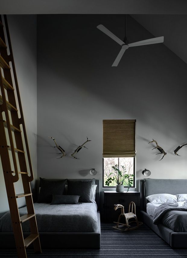 A moody, compact bedroom with two beds arranged along opposite walls, dark grey upholstered bed bases, charcoal striped bedding and a central window with a woven roller blind. A black bedside cabinet, potted plant, wall-mounted reading lights, sculptural branch-like wall decor and a small wooden rocking horse add detail. A tall timber ladder-style feature occupies the left side, while a minimalist three-blade ceiling fan hangs from the dark ceiling. The room has a dark, refined contemporary feel with striped carpet and strong vertical symmetry.