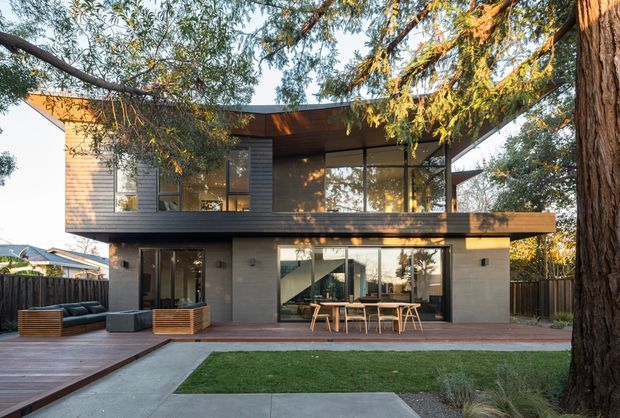 A contemporary two-storey residence viewed from the rear garden, with a projecting upper floor, broad floor-to-ceiling glazing, dark horizontal cladding, concrete lower-level walls, and a sheltered outdoor dining and lounge area. A mature tree frames the facade, while paving, timber decking, and a small lawn create a landscaped courtyard-like setting.