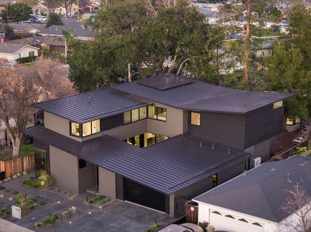 Aerial view of a contemporary multi-level residence in an established suburban neighbourhood. The home has strongly articulated dark metal low-pitched roofs, charcoal horizontal exterior cladding, pale rendered walls, broad dark-framed windows, and an integrated garage. Landscaping and paved paths surround the house, with neighbouring homes and mature trees visible beyond.