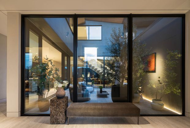 Interior view of a refined contemporary entry or hallway with a large black-framed glazed partition and doorway opening toward a bright double-height living space. A built-in upholstered bench sits in the foreground beside a stone-textured pedestal and potted plants. Warm wall lighting, pale timber flooring, dark frames, and reflected artwork create a calm, sophisticated atmosphere.