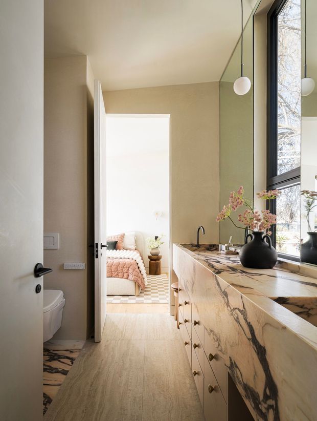 A refined bathroom vanity zone viewed from the bathroom doorway, with a long marble-clad built-in vanity and integrated basin, black tapware, large black-framed window, and spherical pendant lights. A toilet is visible in the foreground, while an open doorway leads directly to a softly furnished bedroom with a bed and bedside table. The space uses warm beige walls, light stone flooring, and dramatic veined marble surfaces with contemporary detailing.