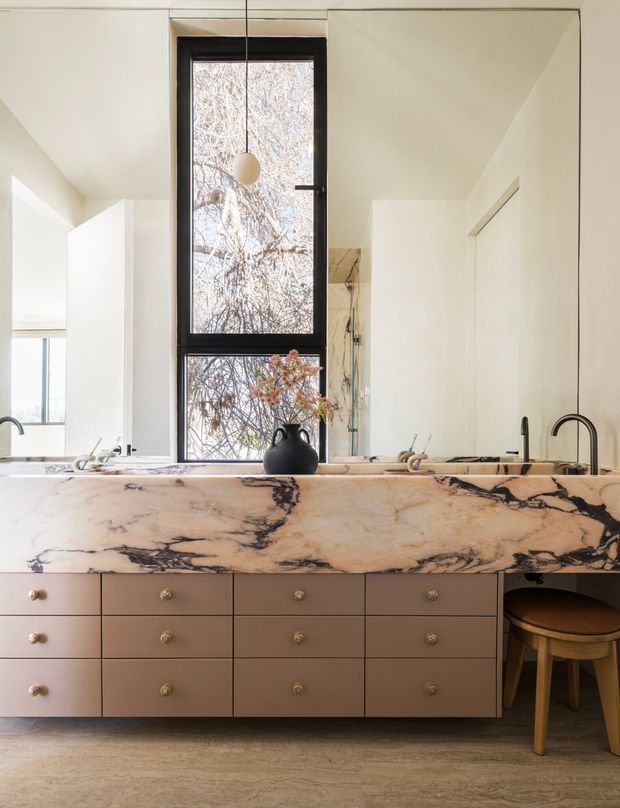 A refined kitchen interior viewed across a large marble island with integrated drawers and brass-toned knobs. The island has dramatic black veining on a warm pale stone surface, with a dark gooseneck tap and a timber stool tucked beneath one end. A tall black-framed window and mirrored surfaces add natural light and depth, while pale walls, timber flooring, and minimal pendant lighting create a warm contemporary setting.