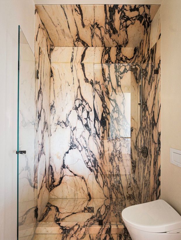 Compact contemporary bathroom featuring a frameless glass walk-in shower fully lined in dramatic veined marble or marble-look stone, including the walls, ceiling, and shower floor. A dark metal shower system and linear drain contrast with the warm cream-and-black stone. A white wall-hung toilet and light neutral wall finish are visible beside the shower.