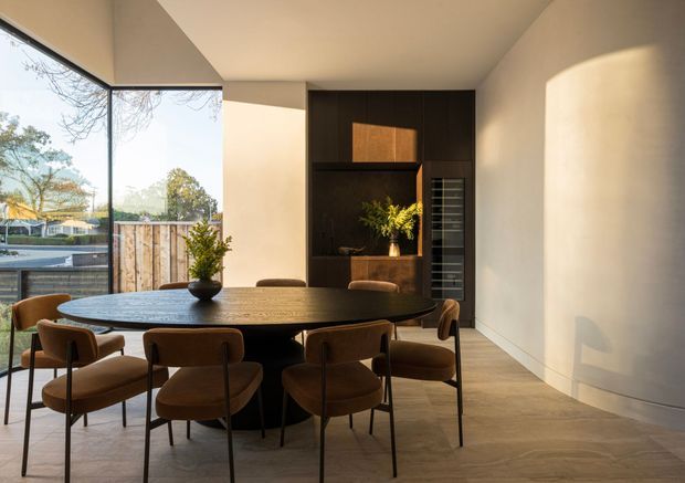 Contemporary dining area with a large round dark dining table and upholstered dining chairs, positioned beside full-height corner glazing. A dark timber built-in feature wall incorporates a recessed fireplace or display niche and vertical wine storage. The room has smooth warm off-white walls, a light stone or concrete-look floor, sculptural curved wall detailing, and warm natural light.