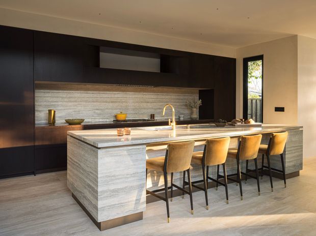 Contemporary kitchen with a large stone island and integrated breakfast bar seating for four. Dark timber cabinetry spans the rear wall, complemented by a horizontally textured stone splashback, pale stone benchtops, timber-toned flooring, and warm brass tapware. A narrow window brings in natural light and greenery, creating a refined, warm and high-contrast interior.