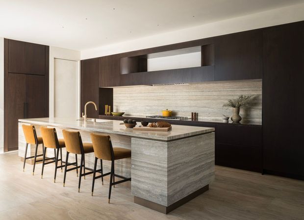 A refined contemporary kitchen centered on a large stone island with seating for five. The space features dark timber cabinetry, a full-height horizontally veined stone splashback, integrated appliances, a brass-toned tap, light timber flooring, and warm tan upholstered bar stools. Minimal recessed ceiling lighting and uncluttered joinery create a sophisticated, balanced interior.