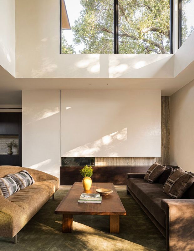 A warm, refined living room with a large brown sectional sofa and matching curved armchair arranged around a dark timber coffee table. The room features a minimalist cream built-in media wall with dark wood and textured stone-like detailing, tall black-framed windows, and a dramatic double-height ceiling opening that brings in abundant natural light and views of trees.