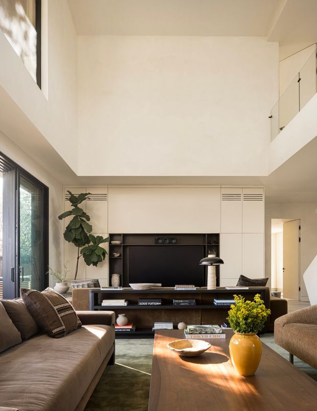 A refined contemporary living room with a double-height void, warm neutral finishes, and abundant natural light. The room features a large upholstered sofa, low timber coffee table, built-in media wall and shelving, a dark stone or timber-toned console, pendant lighting, and tall glazing opening toward greenery. An upper-level balustrade is visible along the right side.