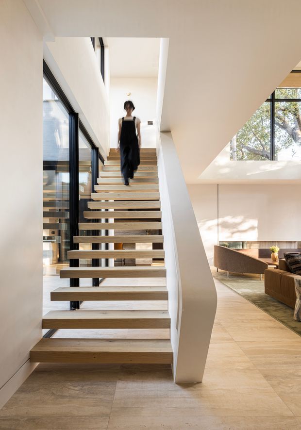 Bright contemporary interior centered on an open-riser staircase with thick light timber treads and a sculptural white side wall. Black-framed floor-to-ceiling glazing runs alongside the stairs, while a partially visible living room with curved seating and large windows appears to the right. Pale stone or tile flooring, white walls, and abundant natural light create an airy, refined atmosphere.