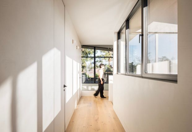 A bright, minimalist residential hallway with pale timber flooring, smooth white walls, flush white doors, and large black-framed windows. Strong natural sunlight creates geometric shadows across the walls and floor, with a person visible near the glazed end of the corridor and views of greenery outside.