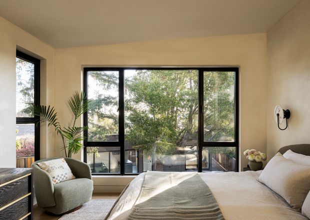 A bright, calm bedroom with a large bed facing expansive black-framed windows, a soft upholstered armchair, indoor palm, bedside furniture, and warm cream walls. The glazing overlooks leafy trees and an outdoor deck or balcony. The room has a simple contemporary fit-out with neutral bedding and natural textures.