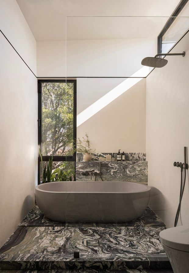 A refined contemporary bathroom featuring a freestanding oval bathtub set on a raised platform clad in dramatic veined natural stone. Matching stone covers the floor and lower wall, while the upper walls and ceiling are white and minimal. A frameless glass shower enclosure with a rain shower occupies the right side, and a tall black-framed window brings in greenery and strong natural light. Matte black fixtures and restrained detailing create a luxurious, sculptural atmosphere.