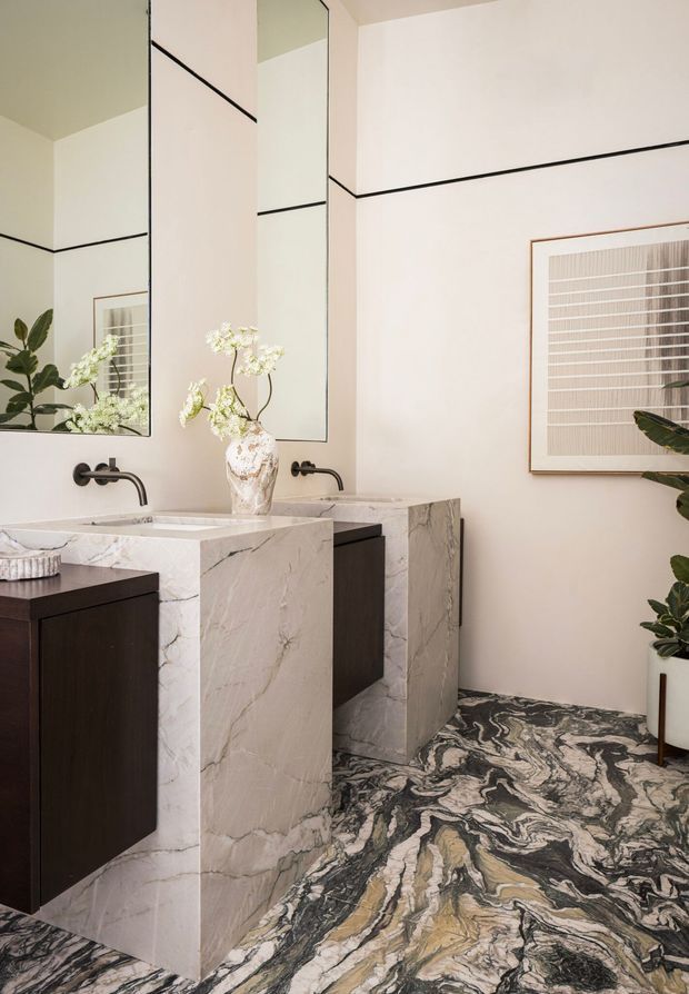 A refined bathroom featuring a large double vanity with sculptural natural-stone surrounds and integrated basins, dark timber cabinetry, wall-mounted dark metal taps, oversized frameless mirrors, and dramatic black-and-white veined stone flooring. Neutral walls, framed artwork, and indoor plants add a calm, luxurious finish.