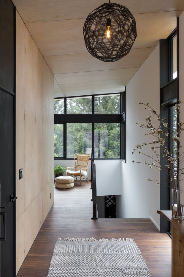A refined residential hallway or landing with dark timber flooring, pale timber wall and ceiling lining, black-framed windows and doors, and a stair opening with black balustrade. The corridor leads toward a bright sitting nook furnished with a woven armchair and ottomans. A sculptural woven pendant light, patterned runner, and tall branch arrangement add warmth and texture. The view through the windows shows leafy greenery outside.