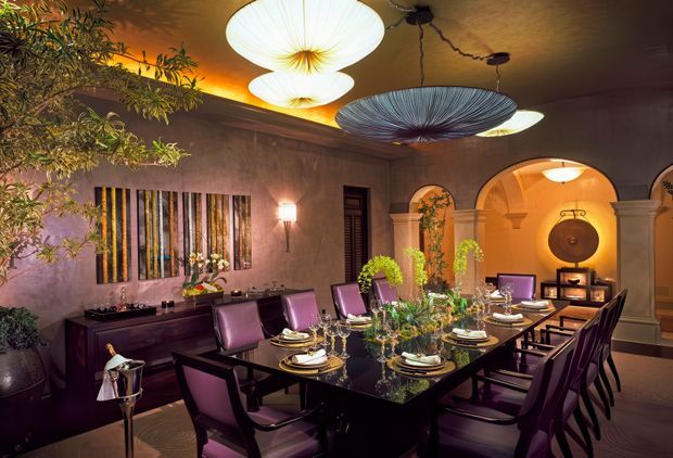 A dramatic, richly decorated dining room with a long dark rectangular dining table set for a formal meal. Deep purple upholstered dining chairs surround the table, which is styled with reflective glassware, plates, candles, and a central floral arrangement. The room features warm textured plaster walls, dark built-in cabinetry, an abstract vertical wall artwork, arched architectural openings, classical columns and mouldings, and oversized sculptural pendant lights. Warm amber lighting creates an intimate, luxurious atmosphere.