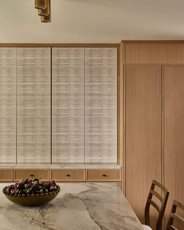 Warm, refined kitchen interior featuring full-height textured light cabinetry, natural wood panelled cabinetry and doors, a pale veined stone benchtop and island, and brass cylindrical pendant lights. A timber dining chair and bowl of fruit sit in the foreground.