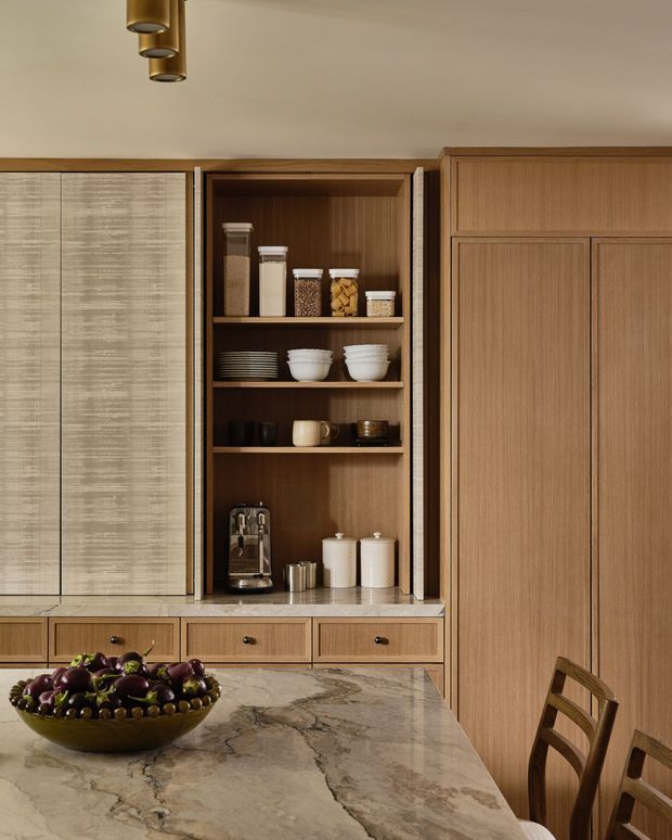 A warm, refined kitchen interior focused on custom timber cabinetry and an open pantry niche. The cabinetry combines vertical wood-grain doors with textured light panels, dark wood shelving, and stone benchtops. Open shelving displays pantry jars, crockery, cups, and a coffee machine, while a large veined stone island occupies the foreground with a bowl of fruit and timber dining chairs. A small brass-toned pendant cluster hangs overhead.