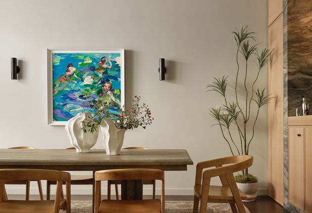 Warm, refined dining area with a solid timber dining table and matching wooden chairs, a colourful framed artwork, sculptural ceramic vases, wall sconces, and a tall indoor plant. A built-in timber-toned joinery wall is visible at right, contributing to the room's crafted, contemporary character.