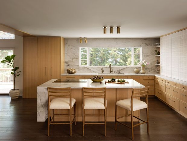 A warm, refined kitchen with extensive light timber cabinetry, a large central island with three stools, and a dramatic full-height marble backsplash and benchtop. Integrated appliances are concealed within the joinery, while a wide window above the sink brings in natural light and views of greenery. The room has dark timber flooring, brass-toned lighting and hardware, and a softly neutral palette.
