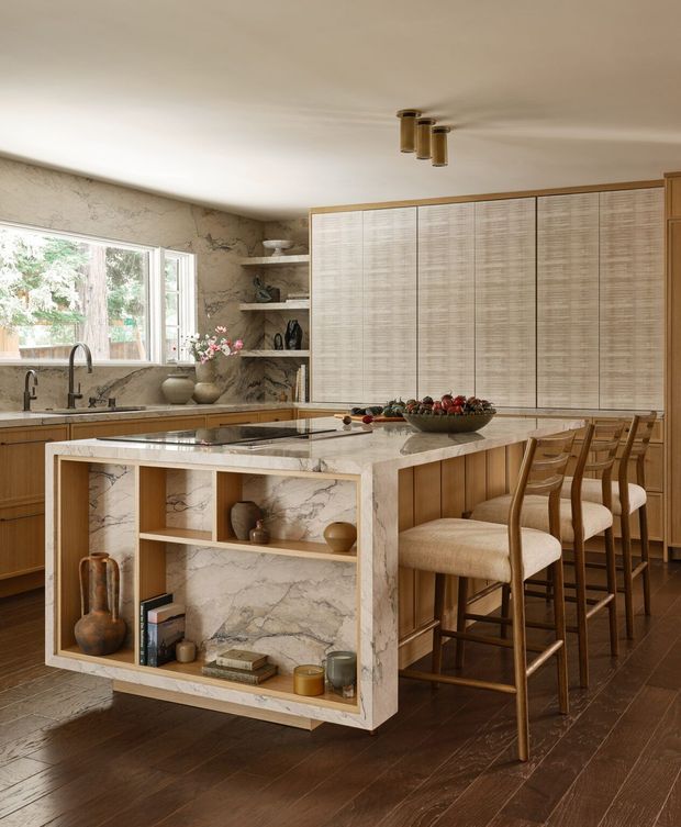 Warm, refined kitchen interior featuring a large central island with a pale veined stone benchtop, integrated open display shelving, timber panelled cabinetry, and seating for several people. The rear wall has tall woven-texture cabinet fronts, while the sink wall includes matching stone surfaces and open shelves. Dark timber flooring, brass-toned pendant lighting, and neutral upholstered bar stools create a sophisticated, natural atmosphere.
