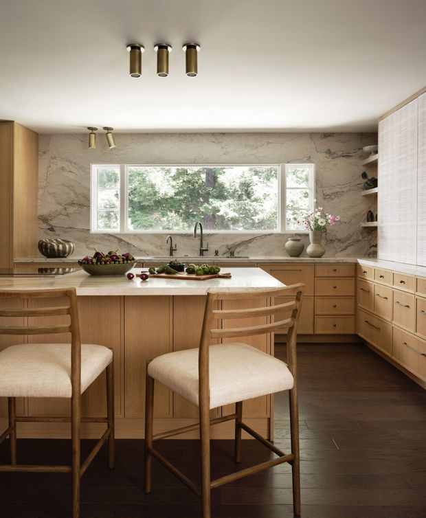 Warm, refined kitchen interior centred on a timber island with upholstered bar stools. The room features extensive light timber cabinetry, a pale veined stone backsplash and benchtop, dark timber flooring, white ceilings, brass-toned cylindrical lighting, and a wide multi-pane window overlooking greenery. Open shelving and decorative objects add a lived-in yet considered finish.