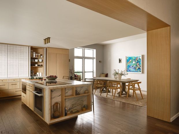 A warm open-plan kitchen and dining area with extensive light timber cabinetry, a large stone island with integrated seating and display shelving, and a timber-framed opening separating the spaces. The dining area includes a long timber table and chairs on a rug, with large black-framed windows, a colourful artwork, and natural light. The space has a refined contemporary character with warm, natural finishes.