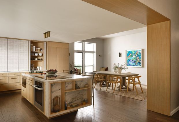 Open-plan kitchen and dining area with warm timber cabinetry, a large central island featuring a veined stone finish and open display shelving, and integrated appliances. The dining zone includes a timber table and chairs on a neutral rug, with large black-framed glazing, white walls, timber flooring, and colourful wall artwork. A substantial timber-framed opening visually connects the spaces.