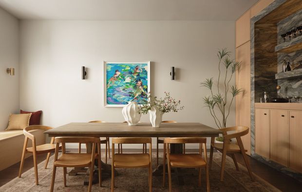 Warm, refined dining room with a long timber dining table and matching wooden chairs, set against pale walls. An abstract colourful artwork hangs above the table, with sculptural ceramic vases and delicate branches as centrepieces. A built-in timber joinery unit with open shelving and dark stone-like backing occupies the right side, while a partial lounge chair and cushion are visible at left. Wall sconces provide understated lighting, and a patterned rug anchors the dining setting.