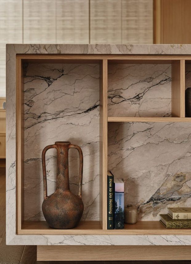 Close-up of a built-in timber display shelving unit framed and backed with richly veined beige natural stone. The open shelves contain a sculptural rust-toned ceramic vessel, a few books, and a small jar. A matching stone slab forms the surrounding frame and lower ledge, creating a warm, refined material-focused interior detail.